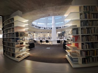 Zdjęcie przedstawia wnętrze ostatniego piętra biblioteki na planie koła. Książki poustawiane na półkach po kole. Wewnątrz koła znajduje się okrągłe okno dachowe.. Kliknij, aby powiększyć zdjęcie.