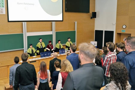 Kadry z inauguracji roku akademickiego w Audytorium Aleksandra Jabłońskiego - kolegium dziekańskie wraz z prorektorką za stołem oraz studenci i nauczyciele oraz administracja siedzący na sali. Kliknij, aby powiększyć zdjęcie.