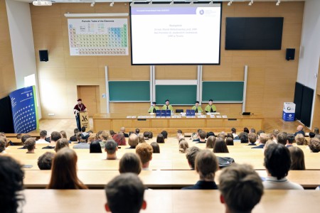 Kadry z inauguracji roku akademickiego w Audytorium Aleksandra Jabłońskiego - kolegium dziekańskie wraz z prorektorką za stołem oraz studenci i nauczyciele oraz administracja siedzący na sali. Kliknij, aby powiększyć zdjęcie.