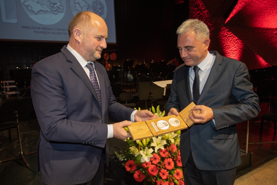 Na scenie sali koncertowej na toruńskich Jordankach stojący po prawej stronie profesor Jarosław Kłaczkow odbiera z rąk stojącego po lewej stronie Marszałka Województwa Piotra Całbeckiego pamiątkowy medal (stylizowane na liczbę sto metalowe odlewy umieszczone w trójdzielnej drewnianej podstawie). Obaj panowie ubrani na galowo, w tle kosz z bukietem kwiatów, puste krzesła i pulpity dla orkiestry.