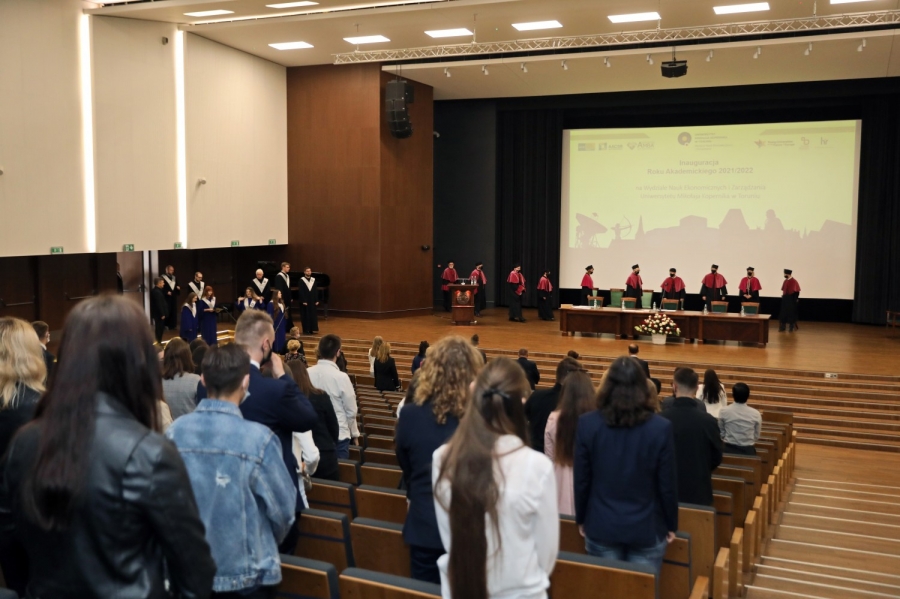 Rzut ogólny na Aulę UMK w trakcie inauguracji roku akademickiego 2021/2022 dla studentów stacjonarnych. Ludzie stojący w trakcie wejścia pracowników Wydziału na scenę. 