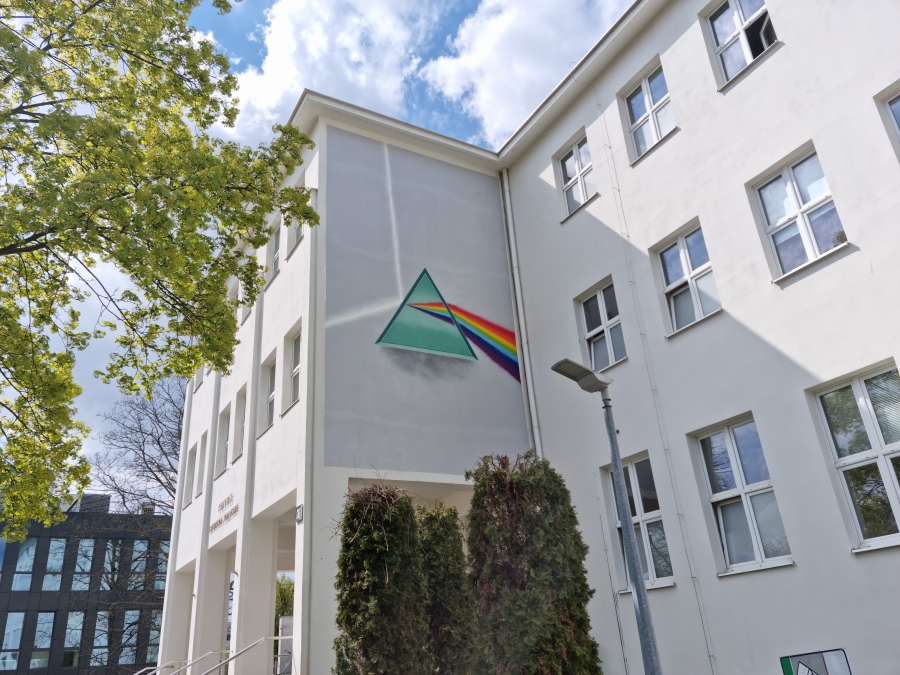 Biała ściana budynku, na której znajduje się duży mural przedstawiający pryzmat rozszepiający światło białe na barwy podstawowe. 