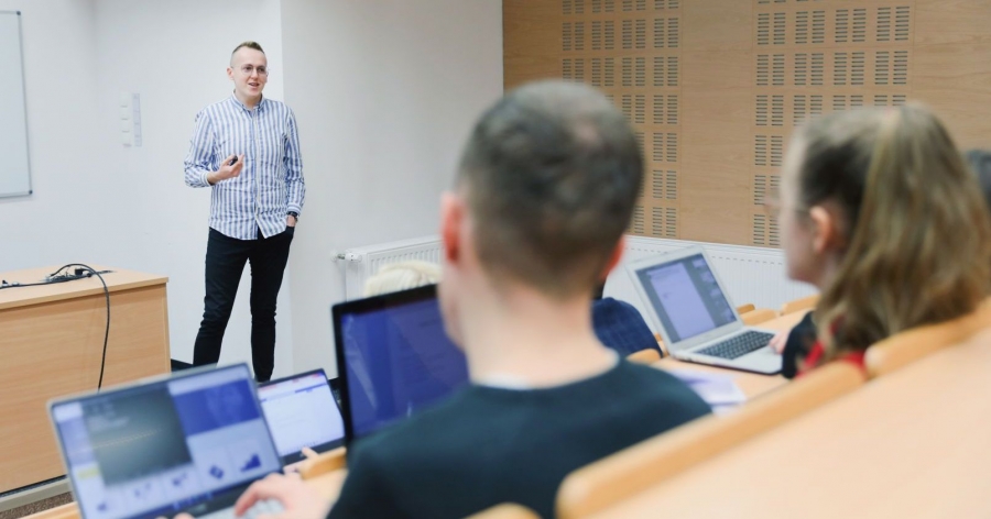 Wykładowca prowadzi zajęcia ze studentami, studenci siedzą tyłem do fotografa i mają włączone komputery