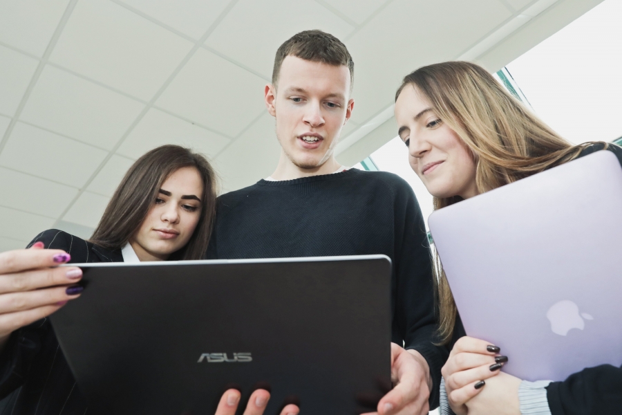 Trójka studentów: 2 kobiety i jeden mężczyzna, stojący obok siebie i patrzący w ekrany dwóch laptopów - zdjęcie podglądowe. 
