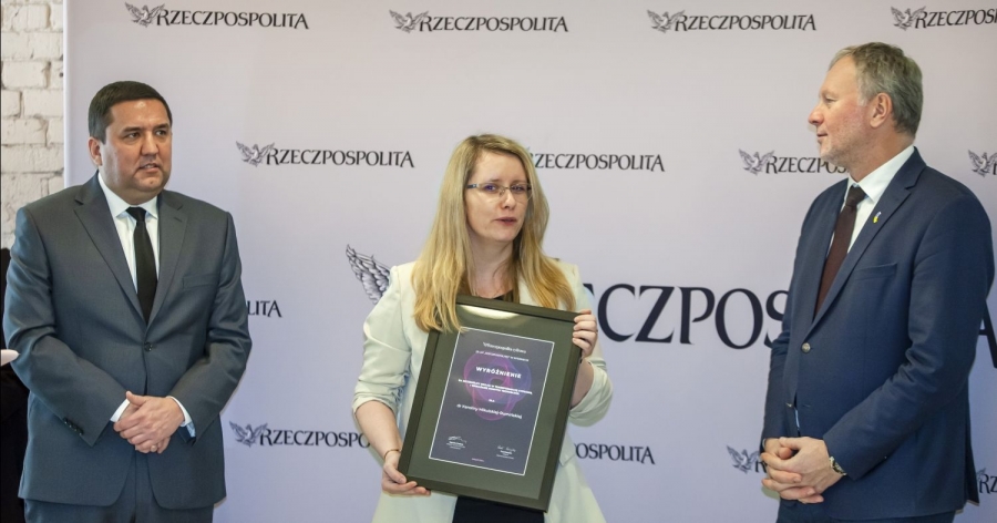 Na środku stoi dr Karolina Mikulska-Rumińska z dyplomem oprawionym w ramie. Po prawej i po lewej stronie stoją mężczyźni w garniturach. Za nimi stoi ścianka z logo gazety 