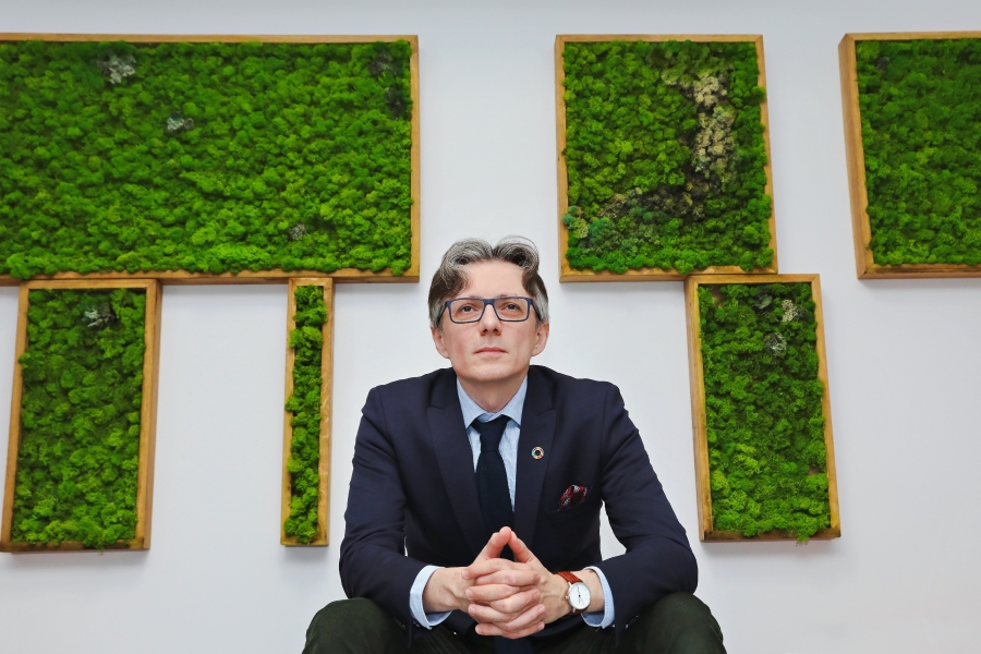 Portret bohatera wywiadu patrzącego przed siebie, siedzącego naprzeciw nas; jest ubrany elegancko (marynarka, koszula, krawat, zegarek); tuż za nim biała ściana z ujętymi w drewniane ramy zielonymi kompozycjami mchu.
