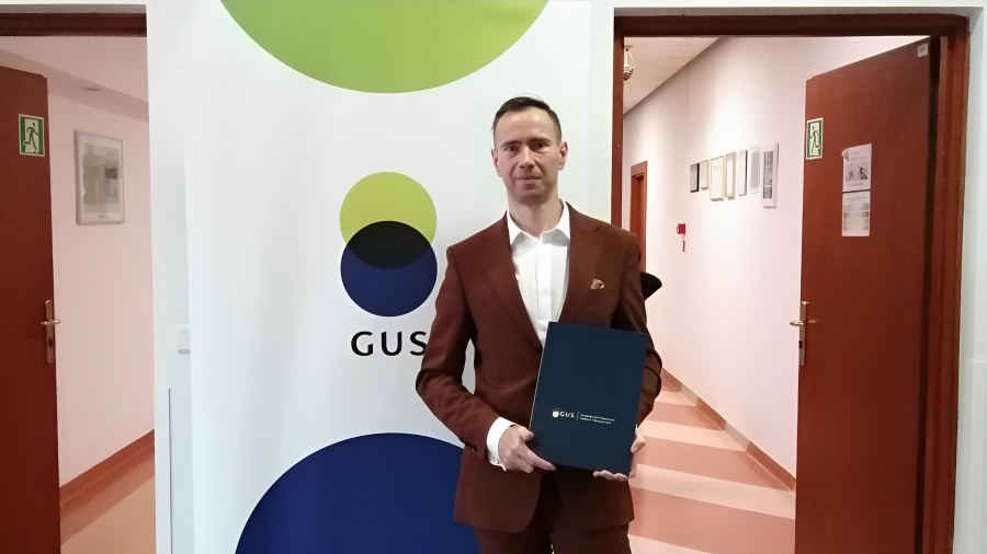 Dr Mateusz Jankiewicz w siedzibie Głównego Urzędu Statystycznego z nagrodą za najlepszą pracę doktorską (teczką). 