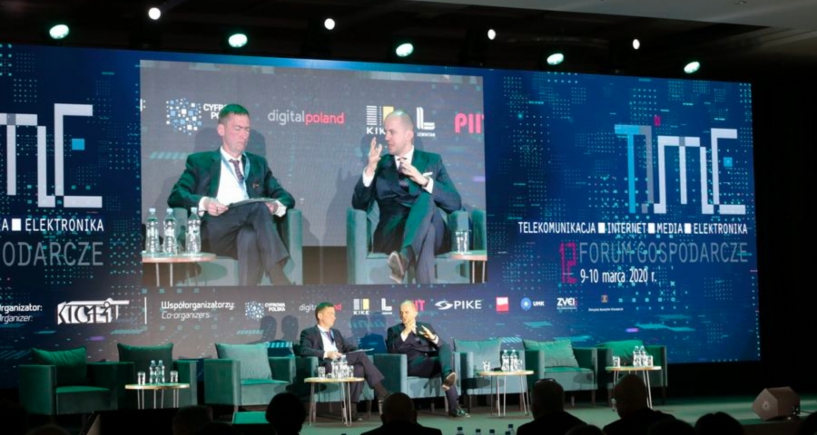 Kadr na scenę konferencyjną. Z tyłu telebim, na których widać dwóch mężczyzn w powiększeniu siedzących na scenie. 