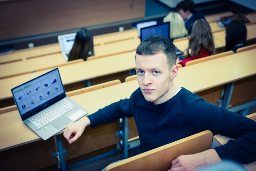Student siedzący w sali audytoryjnej ze wzrokiem skierowanym do obiektywu. W tle inni uczestnicy wykładu. Zdjęcie dekoracyjne.