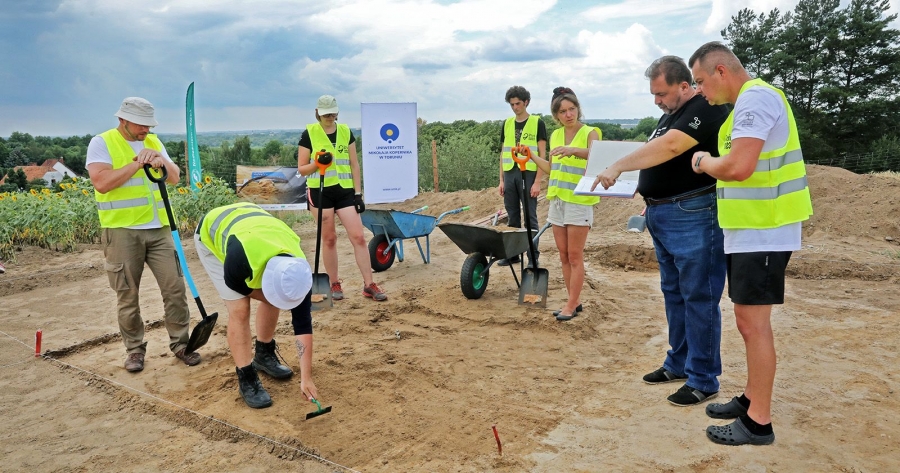 Grupa osób w kamizelkach odblaskowych na wykopaliskach. W tle stoją dwie taczki. Z przodu jedna osoba jest pochylona i trzyma małą gracę, a obok niej stoi osoba i trzyma łopatę