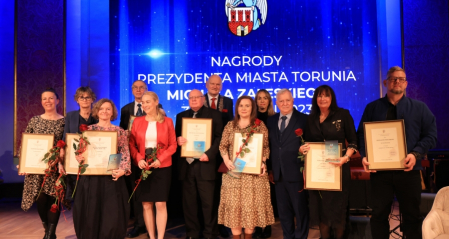 Grupa nagrodzonych stoi razem i w ręku trzyma dyplomy. W tle wyświetla się slajd prezentacji z nazwiskami nagrodzonych oraz zdjęciem awersu i rewersu medalu Thorunium