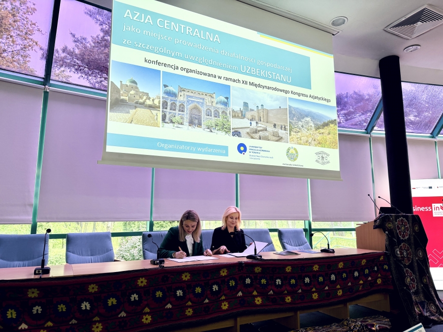 Dwie kobiety siedzą przy stole konferencyjnym i podpisują dokumenty podczas wydarzenia poświęconego Azji Centralnej, ze szczególnym uwzględnieniem Uzbekistanu. Za nimi widoczny jest duży ekran z prezentacją o treści: ?Azja Centralna jako miejsce prowadzenia działalności gospodarczej ze szczególnym uwzględnieniem Uzbekistanu?, w ramach XII Międzynarodowego Kongresu Azjatyckiego. Na ekranie znajdują się także zdjęcia z Uzbekistanu oraz logotypy organizatorów, w tym Uniwersytetu Ekonomicznego we Wrocławiu i Ambasady Uzbekistanu.