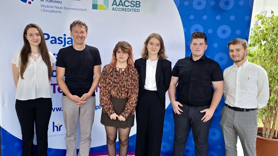 Grupa sześciu osób stoi na tle ścianki z logotypami Uniwersytetu Mikołaja Kopernika w Toruniu i akredytacji AACSB. Od lewej: kobieta w białej bluzce i czarnych spodniach, mężczyzna w czarnym t-shircie i jasnych spodniach, kobieta w wzorzystej bluzce i ciemnej spódnicy, kobieta w czarnej marynarce i białej koszuli, mężczyzna w czarnej koszuli i spodniach oraz mężczyzna w białej koszuli i szarych spodniach. W tle roślina w doniczce