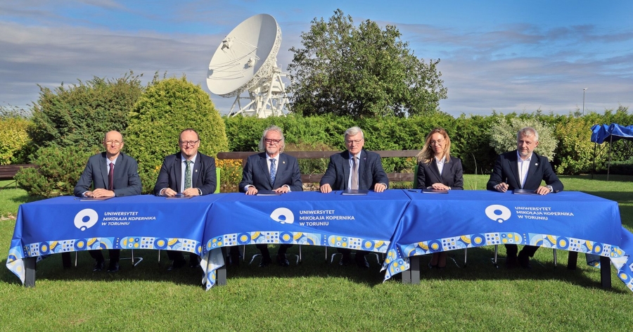 Five men and one woman sit at a table set outdoors, with a radio telescope in the background. They look into the camera lens.