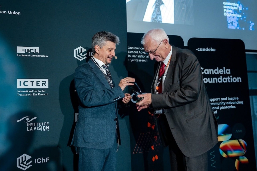 Zdjęcie przedstawia scenę z konferencji naukowej lub medycznej, na której prof. dr hab. Maciej Wojtkowski wręcza prof. dr. hab. Andrzejowi Kowalczykowi statuetkę. Prof. dr hab. M. Wojtkowski stoi przodem do prof. dr. hab. A. Kowalczyka i lekko bokiem do obiektywu. Jest to mężczyzna w średnim wieku, z krótkimi, szpakowatymi włosami. Ubrany jest w elegancki, ciemnogranatowy garnitur i krawat. Na szyi ma zawieszoną smycz identyfikacyjną. Właśnie przekazuje statuetkę do rąk drugiego mężczyzny. Uśmiecha się. Prof. dr hab. A. Kowalczyk jest starszy, ma siwe włosy i okulary. Ubrany jest w ciemny, białą koszulę i czerwony krawat. Na szyi ma również zawieszoną smycz z identyfikatorem. Pochyla się nieco do przodu, by przyjąć nagrodę. Uśmiecha się łagodnie. Obaj mężczyźni trzymają w dłoniach małą, przezroczystą statuetkę, która wygląda na wykonaną ze szkła lub akrylu. Scena rozgrywa się na tle ciemnej, profesjonalnej ściany wystawowej, pełnej logotypów i napisów związanych z konferencją. Zdjęcie ma uroczysty i pozytywny charakter, uwieczniając moment wręczenia prestiżowej nagrody podczas międzynarodowej konferencji naukowej. Obaj mężczyźni wydają się zadowoleni i honorowi.