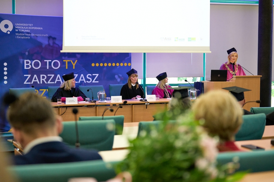 Nauczyciele akademiccy w togach i biretach siedzący w eleganckiej sali.