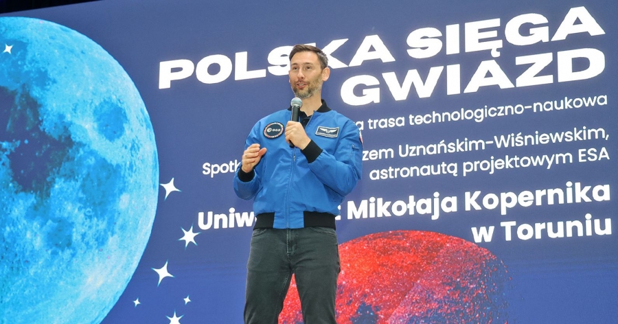Astronaut Sławosz Uznański-Wiśniewski stands with a microphone in front of the inscription 
