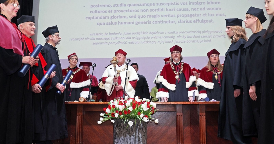 Ceremonia promocji doktorskiej, kolegium rektorskie stoi za stołem prezydialnym, po bokach promotorzy i nowo promowani doktorzy. W tle ekran z treścią ślubowania