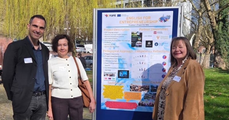 Zdjęcie wykonane w przestrzeni parku. Na pierwszym planie stand z dużym plakatem konferencyjnym. Obok plakatu stoją trzy uśmiechnięte osoby, uczestnicy projektu  ?English for Entrepreneurship?.
