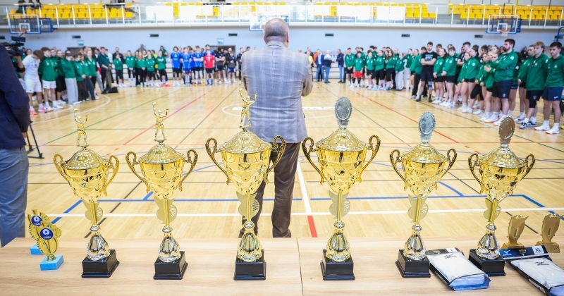 Uroczystość wręczenia nagród w hali sportowej. Na pierwszym planie rząd sześciu złotych pucharów ustawionych na stole. W tle grupy młodych sportowców w strojach treningowych oraz prowadzący ceremonię.