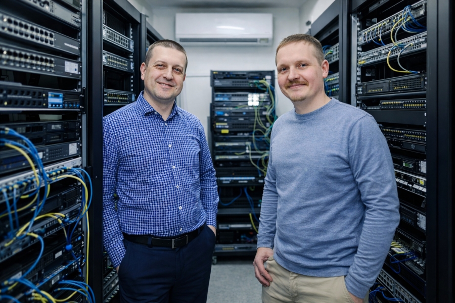 Dwóch uśmiechniętych specjalistów IT (prof. Piotr Masłowski i prof. Mariusz Piwiński) stojących w serwerowni między szafami ze sprzętem sieciowym.
