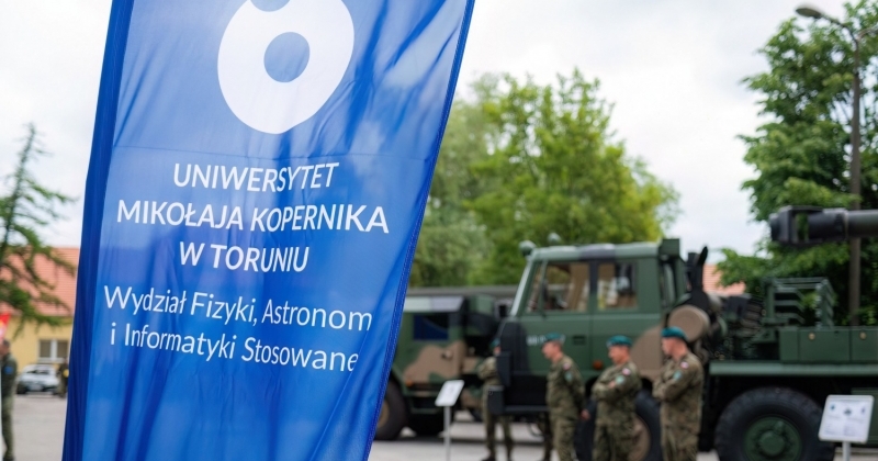 Flaga niebiesko-biała z logo i podpisem WFAiIS oraz Uniwersytetu Mikołaja Kopernika w Toruniu w tle pojazd wojskowy i żołnierze