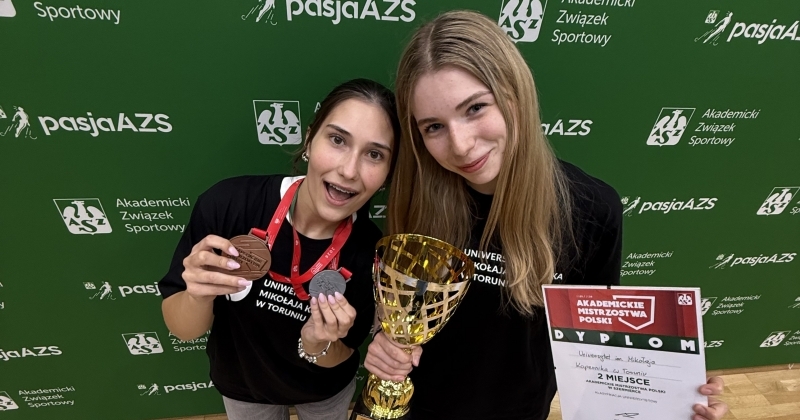 Oto podpis pod zmieniacz (slider) dostosowany do zdjęcia (ponieważ są na nim zawodniczki, a nie Jan Jurkiewicz) oraz tekst alternatywny dla osób słabowidzących:  **Podpis pod zmieniacz:** AMP: Nadia Jaszczołt i Emilia Szefera ze srebrnym pucharem za wicemistrzostwo drużynowe uniwersytetów.  **Tekst alternatywny (ALT):** Dwie uśmiechnięte reprezentantki UMK Toruń w czarnych koszulkach uniwersyteckich prezentują zdobyte na Akademickich Mistrzostwach Polski medale, dyplom za drugie miejsce oraz złoty puchar na tle zielonej ścianki z napisami Akademicki Związek Sportowy.
