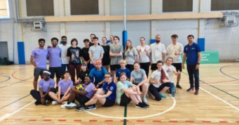 Grupa uczestników i organizator V Uczelnianego Turnieju Quatrokorfballu pozują wspólnie do pamiątkowego zdjęcia na parkiecie hali sportowej UMK, stojąc i siedząc przed żółtym koszem korfballowym.
