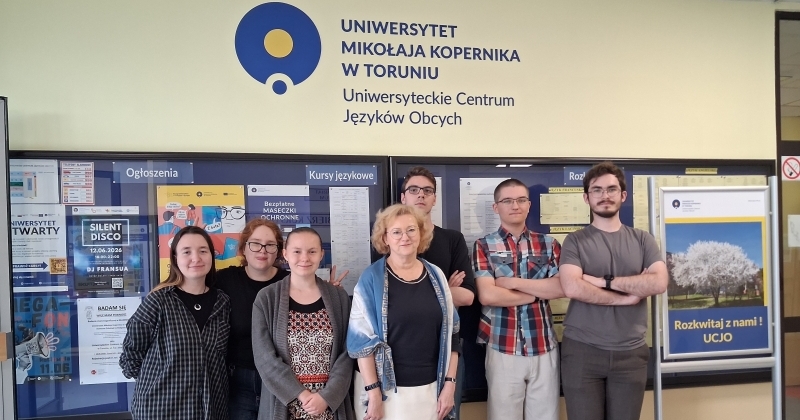 Na planie pierwszym stoją laureaci dwóch konkursów Mistrz Języka Specjalistycznego, które odbyły się 13 maja b.r.na WNH: od lewej ? studentki archiwistyki i zarządzania dokumentacją: Małgorzata Chojnacka, Anna Norkowska i Wiktoria Grochowiecka; pośrodku stoi pani mgr Dorota Hoffmann ( wykładowca Języka angielskiego w UCJO). Dalej po jej prawej stronie stoją studenci: Arkadiusz Dankowski i Wiktor Piasecki, studenci II roku historii oraz Jakub Grubecki ? student II roku archeologii. W tle widoczne są tablice ogłoszeń i nazwa jednostki UMK ? Uniwersyteckie Centrum Języków Obcych. Z prawej strony stoi roll-up promujący UCJO o nazwie ?Rozkwitaj z nami?.