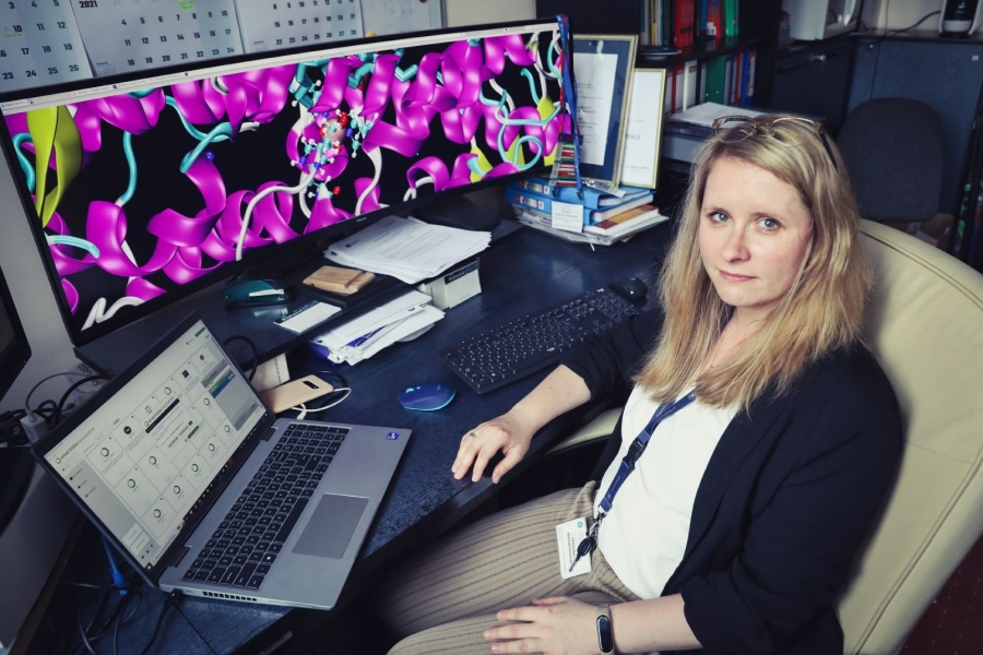 Dr hab. Karolina Mikulska-Rumińska, prof. UMK siedząca przy biurku w biurze. Na biurku znajduje się laptop oraz duży monitor wyświetlający kolorowy model 3D struktury białkowej.