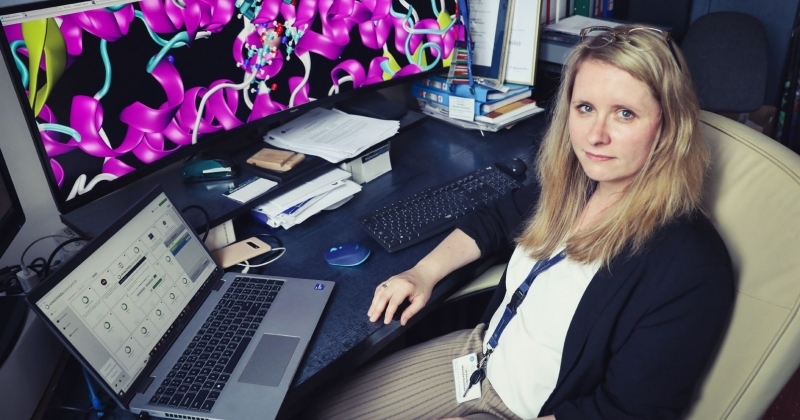 Dr hab. Karolina Mikulska-Rumińska, prof. UMK siedząca przy biurku w biurze. Na biurku znajduje się laptop oraz duży monitor wyświetlający kolorowy model 3D struktury białkowej.