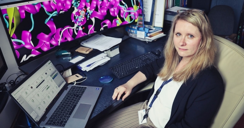 Dr. hab. Karolina Mikulska-Rumińska, NCU Prof., sitting at a desk in an office. On the desk, there is a laptop and a large monitor displaying a colorful 3D model of a protein structure.