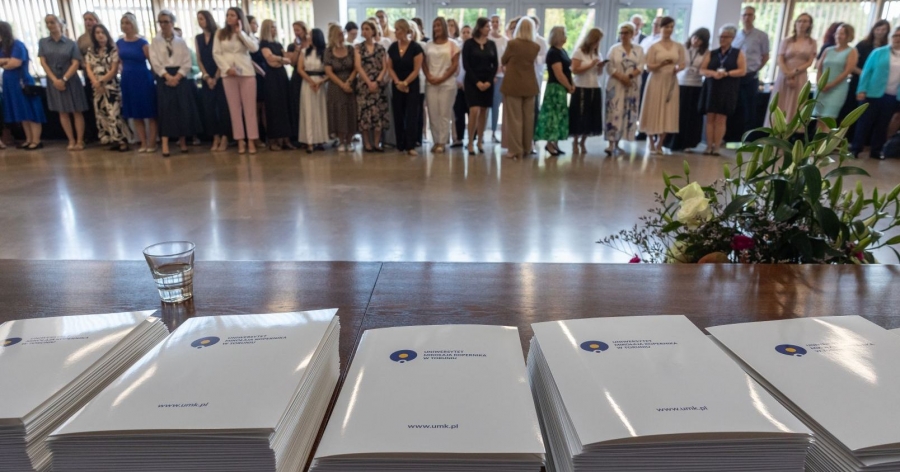 Na pierwszym planie teczki z logiem UMK, w tle grupa osób czekających na odbiór nagrody
