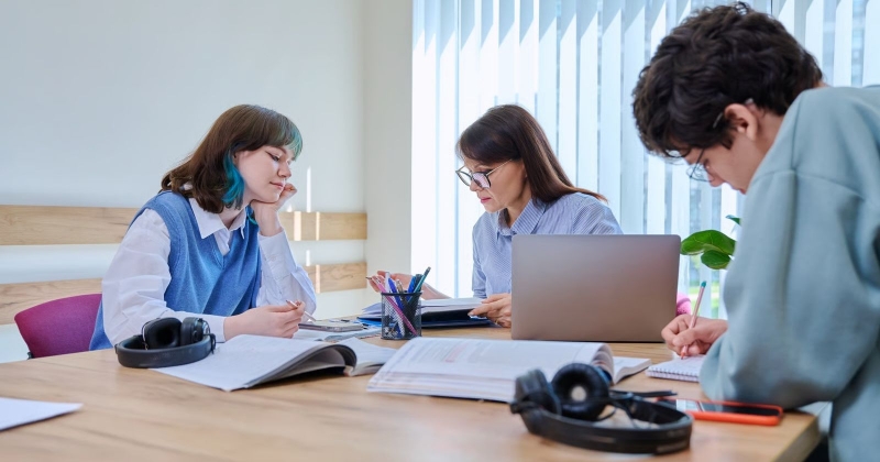 Three people are sitting at a table together; they have open books or notebooks in front of them; two of them are talking to each other, and one is writing something down in a notebook
