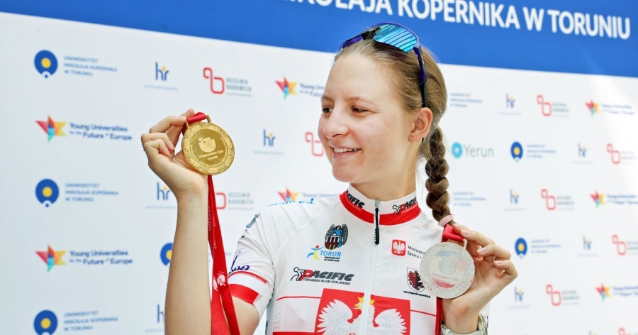 Natalia Krześlak pozuje do zdjęcia z dwoma medalami Akademickich Mistrzostw Świata w kolarstwie, w tle widać ściankę z logo UMK