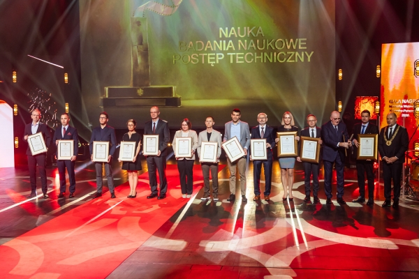 Grupa laureatów stojąca na scenie z oprawionymi w ramy dyplomami podczas Gali Nagród Marszałka Województwa Kujawsko-Pomorskiego w kategorii ?Nauka, badania naukowe, postęp techniczny?.