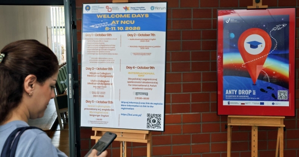Plakat promujący projekt anty drop out i plakat welcome days na sztalugach w holu biblioteki, w ktorym stoi równiez osoba patrzaca w telefon